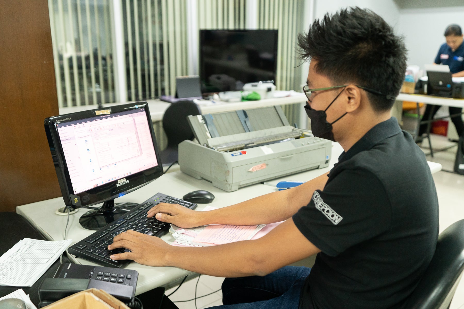 Office Worker wearing a black shirt, glasses and a mask doing data entry using a computer and printers